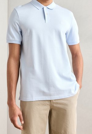 Mand iført lyseblå kortærmet poloshirt og beige shorts, stående med den ene hånd i lommen mod en ensfarvet baggrund.