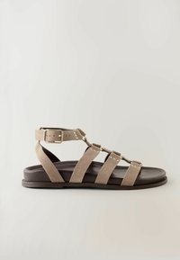 Flache Sandale mit beigen Wildlederriemen, goldfarbenem Schnallenverschluss und schwarzer Sohle, entworfen für den Freizeitgebrauch.