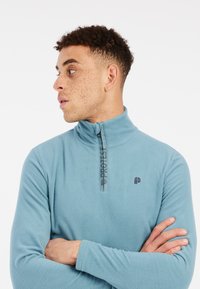 Pullover in pile azzurro chiaro con colletto alto, zip a metà e dettagli del logo neri sulla zip e sul petto. Tessuto morbido, maniche lunghe.