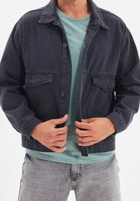 Tmavě modrá džínová bunda se dvěma kapsami na prsou, šedými knoflíky a mírně volným střihem. Nenošena přes světle tyrkysové tričko. Texturovaný povrch látky.