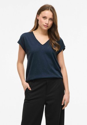 Femme aux longs cheveux bruns portant un haut bleu marine à col en V et manches courtes, ainsi qu'un pantalon noir, posant avec une main dans la poche sur fond blanc.