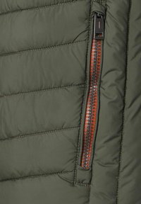 Die olivgrüne isolierte Jacke hat einen gesteppten Stoff. Der Reißverschluss in kontrastierendem Orange verfügt über schwarze Hardware-Details.