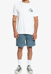 Vit bomulls t-shirt med en svart logga på bröstet, kombinerad med blå kordflorshorts och grå sneakers med vita sulor samt ankelsockar.