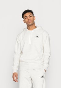 Hoodie gris clair en tissu doux, avec une capuche standard, des cordons de serrage et un logo noir sur la poitrine. La coupe est décontractée.