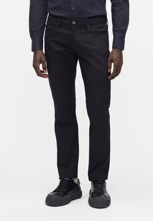 Man draagt zwarte slim-fit broek, zwarte leren sneakers en een donkerblauw overhemd met lange mouwen, staande tegen een witte achtergrond.