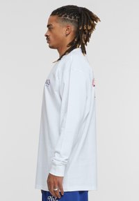 Langärmliges weißes Baumwollhemd mit lockerer Passform. Verfügt über gerippte Bündchen und ein grafisches Design auf der Vorderseite. Getragen mit blauen Mesh-Shorts.