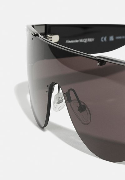 Occhiali da sole con una grande montatura nera curva e lenti scure. Presentano morbidi cuscinetti nasali trasparenti e accenti metallici, mostrando un design moderno.