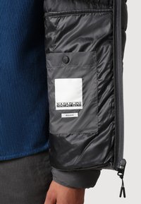Black puffer jacket sleeve with a gray pocket labeled "Napapijri" and "Regular Fit" over a blue ribbed sweater and dark pants.