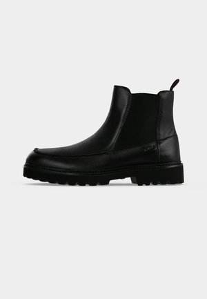 Bottine noire en cuir avec semelle texturée, panneaux latéraux élastiques et languette de tirage sur fond gris clair.