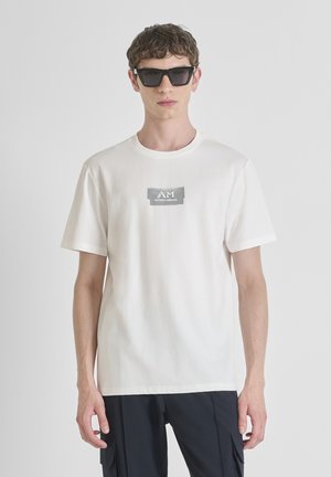 Camiseta de algodón blanca con un logo en el pecho en gris. El modelo la combina con gafas de sol rectangulares negras. Diseño sencillo, de mangas cortas.