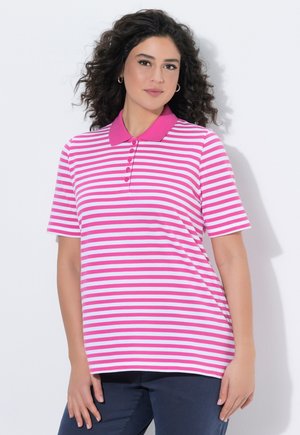 Kvinde med krøllet mørkt hår iført en lyserød og hvidstribet kortærmet polo og mørke bukser, der står mod en hvid baggrund.