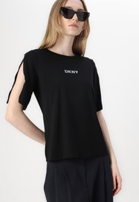 Juodas DKNY trumpomis rankovėmis marškinėliai, pagaminti iš minkštos medžiagos. Sukurti su išpjovimais ant rankovių ir sidabriniu logotipu krūtinėje.