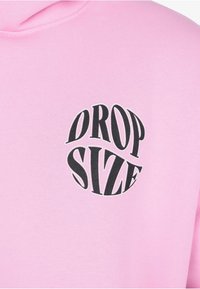 Rosa Stoff mit schwarzem, dickem Text "DROP SIZE" in einer verzerrten, kreisförmigen Form, die auf der Brustpartie eines Kleidungsstücks zentriert ist.