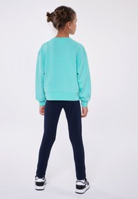 Felpa turchese con una vestibilità comoda e polsini a costine, abbinata a leggings blu navy e sneakers bianche e nere, vista da dietro.