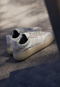 Ljust beigefärgade sneakers med en blandning av slät och texturerad mocka, som har tre sidostreck och en kraftig, krämfärgad sula.