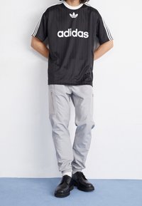 Mustat Adidas t-paita valkoisella logolla, kolmella valkoisella raidalla hihoissa. Vaaleanharmaat housut, joissa on vetoketjut ja joustavat hihnat. Mustat nauhakiinnityksellä varustetut kengät.