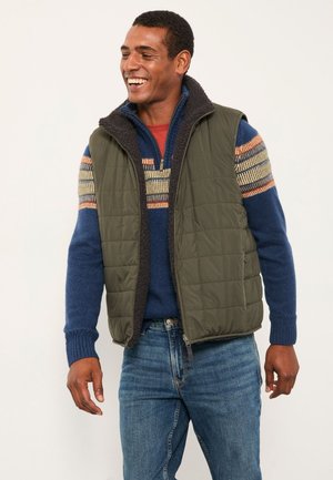 Mann lacht, trägt einen blauen Strickpullover mit gestreiftem Muster, eine olivfarbene gesteppte Weste und blaue Jeans, steht vor einem schlichten Hintergrund.