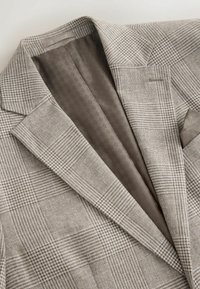 Béžové a hnedé kárované sako s hodvábnou vzorovanou podšívkou a golierom s výrezom, položené na svetlom povrchu.