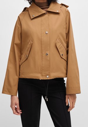 Giacca con cappuccio marrone chiaro con bottoni a pressione e tasche con patta inclinate indossata sopra una maglietta marrone e pantaloni neri.