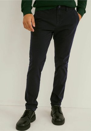 Mężczyzna ubrany w ciemnogranatowe spodnie o dopasowanym kroju, czarne sznurowane buty oraz zielony sweter, stoi z jedną ręką w kieszeni.