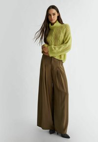 Maglione a collo alto lavorato a maglia verde chiaro con design a treccia abbinato a pantaloni marroni a gamba larga e décolleté nere a punta.