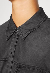 Camicia in denim nero con colletto classico, piastra dei bottoni e dettagli di cucitura visibili sul tessuto. La texture appare liscia.