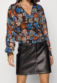 Femme portant un chemisier à manches longues à fleurs bleues, oranges et blanches, associé à une mini-jupe en cuir noire avec des coutures blanches.