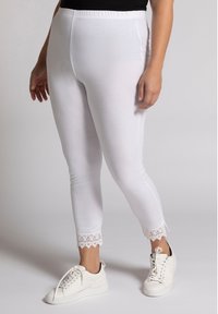 Legging blancs avec une taille élastique, ornés de dentelle au niveau des chevilles. Associés à des baskets blanches, mettant en valeur un look décontracté.
