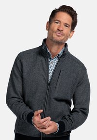 Mann mit braunen Haaren, der eine dunkelgraue Zip-Up-Jacke über einem gemusterten Hemd trägt, vor einem schlichten weißen Hintergrund.