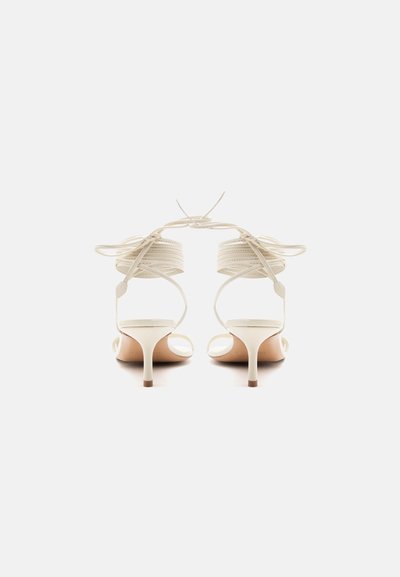 Ζευγάρι λευκών σανδαλιών με ψηλό τακούνι, λουριά που δένουν στον αστράγαλο και ανοιχτά δάχτυλα, φωτογραφημένα από πίσω σε λευκό απλό φόντο.