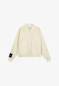 Chaqueta bomber crema con cierre frontal, dos grandes bolsillos frontales, puños elásticos y una etiqueta de tela negra en la manga izquierda. Textura suave.