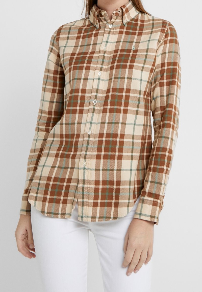 Femme portant une chemise à carreaux beige, marron et bleu clair à boutons avec manches longues et un pantalon blanc sur un fond uni.