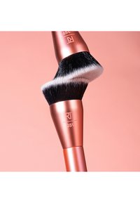 To vinklede makeup-børster med sorte og hvide børstehår og rosaguld håndtag krydser hinanden mod en ferskenfarvet baggrund.
