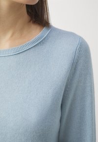 Vaaleansininen villapaita, jossa on ribbireunus ja pehmeä, teksturoitu neule. Lähikuva korostaa sileää kangasta ja olkasauman yksityiskohtia.
