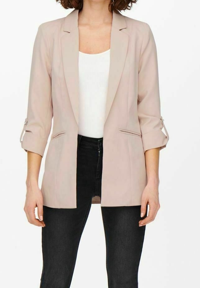 Femme portant un blazer beige clair aux manches retroussées sur un haut blanc et un pantalon noir, debout devant un fond blanc uni.