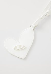 White heart-shaped bag charm made of faux leather, featuring stitched edges and a logo accent in the center. Adjustable strap included.