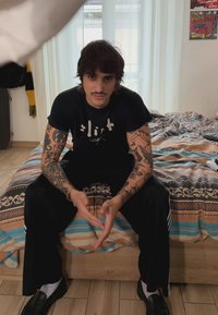 Must Black T-särk valge teksti, mustad püksid, detailidega tätoveeringud kätevartel, heledal põrandal, mustriga voodipesu, pehme valgus suurtest akendest.