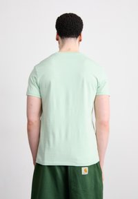 Tricou scurt verde deschis, cu un guler rotund, realizat din țesătură moale. Portat cu pantaloni scurți verzi închisi, cu un patch cu logo. Vedere din spate.