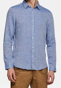 Svetle modrá košile s knoflíky vyrobená z texturované látky, obsahující límec, dlouhé rukávy a bílé knoflíky, v kombinaci s khaki kalhotami.