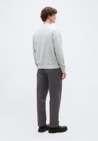 Νέος άνδρας με σγουρά μαλλιά που στέκεται, στραμμένος προς τα δεξιά, φοράει ανοιχτό γκρι φούτερ, σκούρο γκρι παντελόνι και μαύρα παπούτσια σε απλό φόντο.