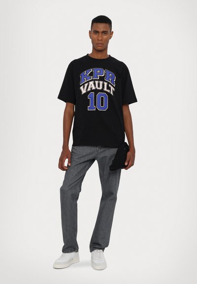 T-shirt nera con grafica blu e bianca "KPR VAULT 10", abbinata a jeans grigi. Il modello tiene un cappellino nero. Scarpe bianche completano l'outfit.