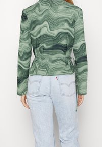 Blazer à motif marbré vert avec manches longues, taille ceinturée et tissu lisse. Associé à un jean en denim bleu clair.