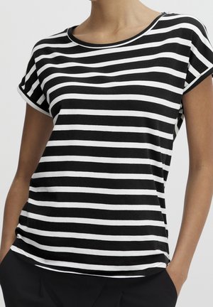 Chemise à manches courtes à rayures horizontales noires et blanches portée par une personne les mains dans les poches, fond clair uni.