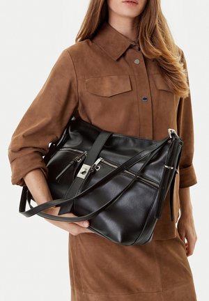 Frau trägt ein braunes Wildleder-Hemdkleid und hält eine große schwarze Lederhandtasche mit silbernem Reißverschluss und Schnallendetails.