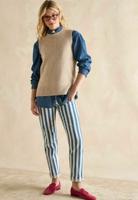 Beige gestrickter ärmelloser Pullover über einem Jeanshemd, kombiniert mit blau-weiß gestreiften Hosen und rosa Mokassins. Flacher Saum und umgeschlagene Bündchen.