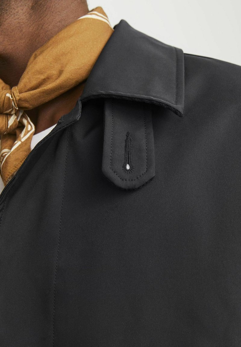 Gros plan d'une personne portant un manteau noir avec des détails d'épaulettes et une écharpe marron à motif nouée autour du cou.