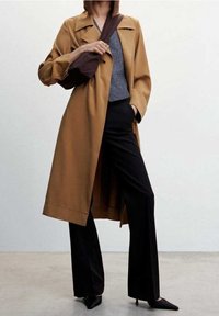 Long brown coat made of smooth fabric, featuring a notched collar and wide sleeves, paired with a gray top and black trousers, accentuated with pointed shoes.