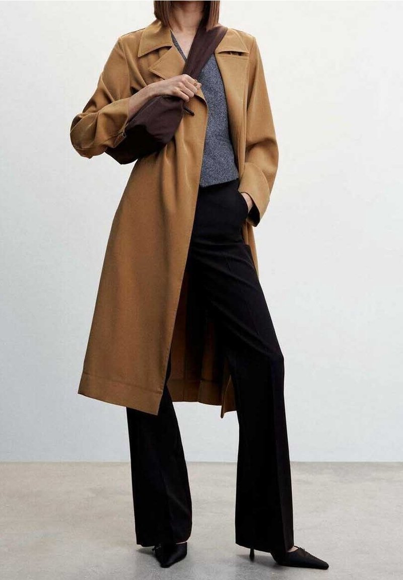 Long brown coat made of smooth fabric, featuring a notched collar and wide sleeves, paired with a gray top and black trousers, accentuated with pointed shoes.