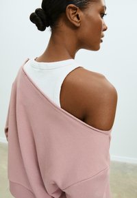 Felpa oversized rosa chiaro con design della spalla scoperta, indossata sopra una canotta bianca. Materiale in misto cotone; trama sottile visibile.
