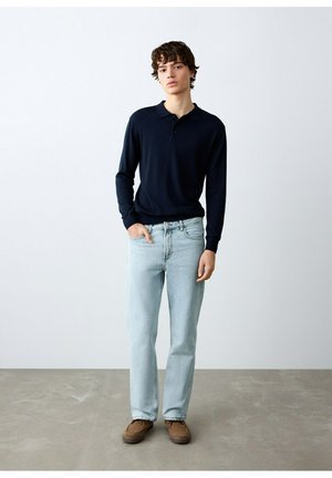 Mladi moški stoji oblečen v črn polo z dolgimi rokavi, svetlo modre kavbojke in rjave čevlje, ob običajni sivi steni in tleh.